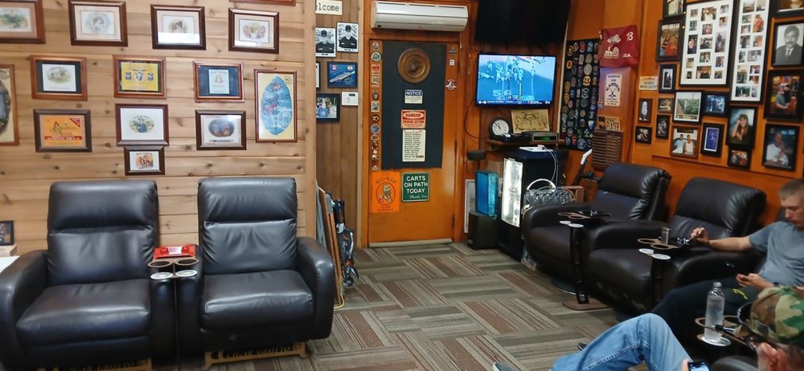 The Cigar Grotto lounge with black leather recliners, framed cigar art and sports on TV